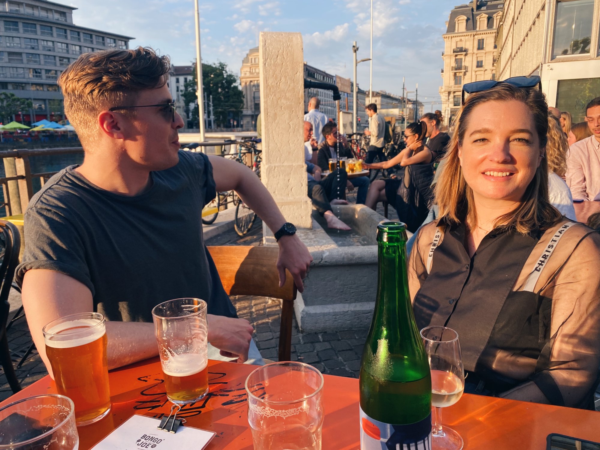 Bongo Joe at golden hour — people at tables, green wine bottle, the Rhône behind