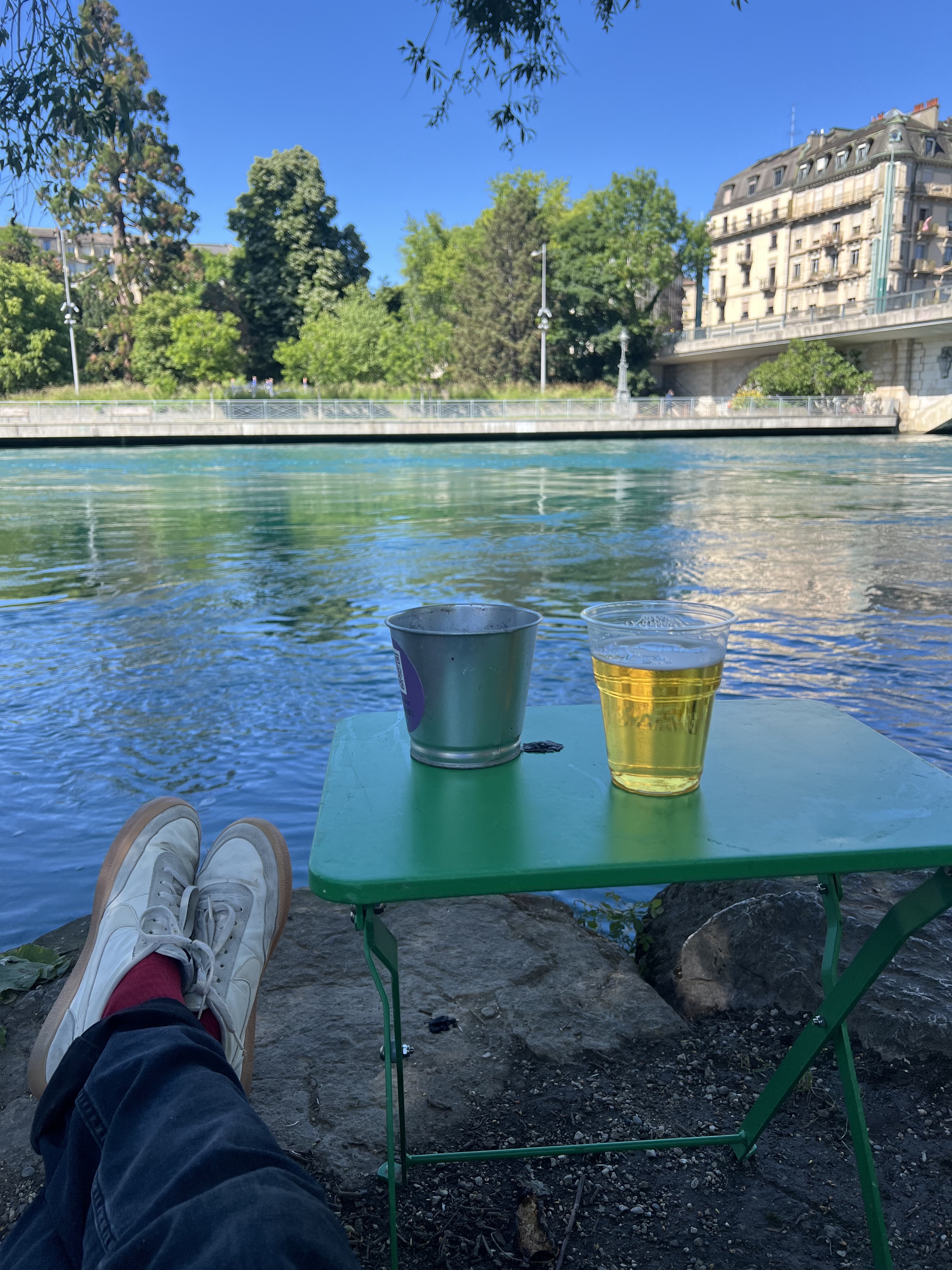 A plastic cup of cold beer on a green table by the Rhône river at La Barje Geneva, white sneakers in the foreground