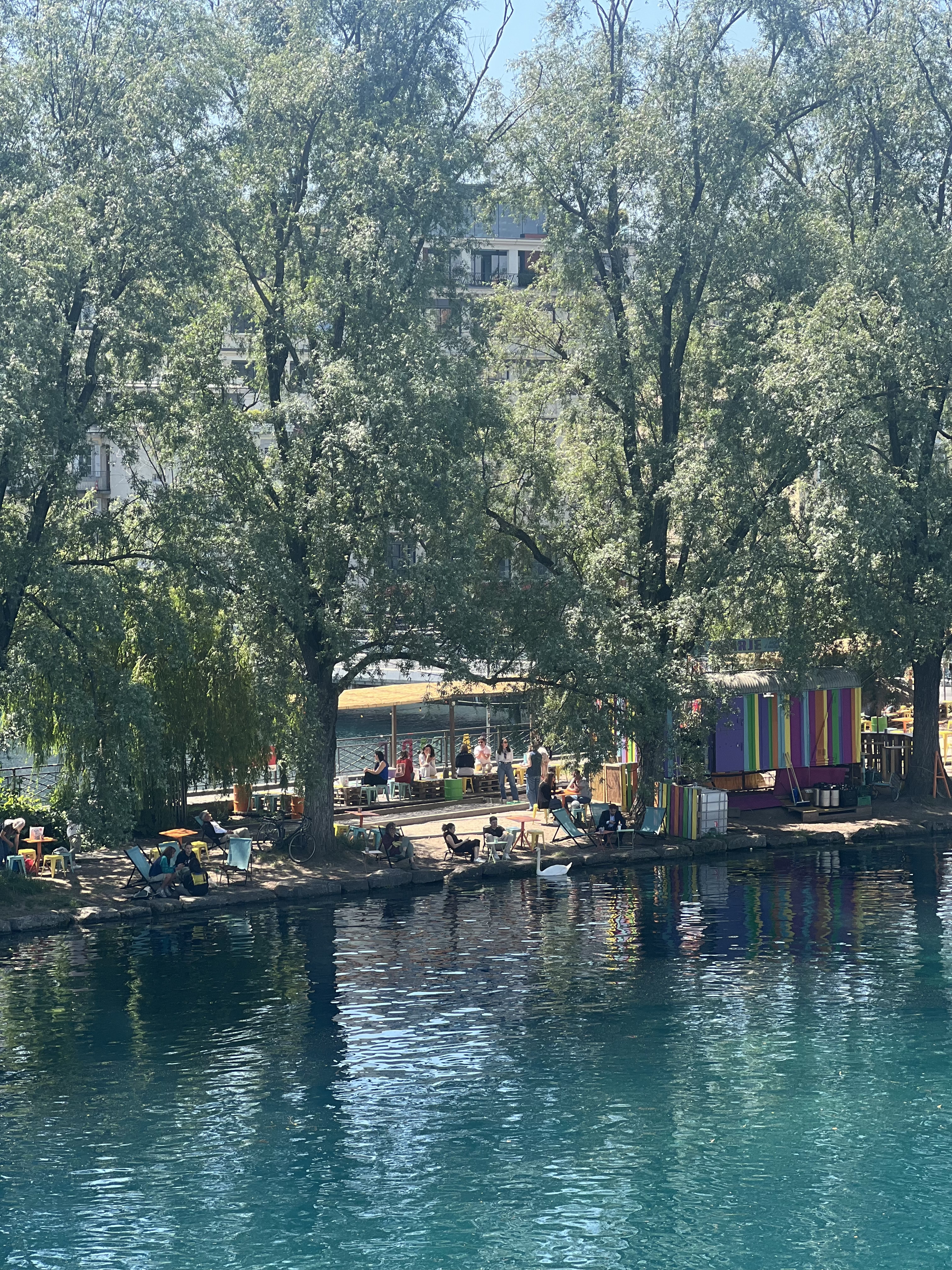 Wide view of La Barje Geneva — colourful bar shed, willow trees, deck chairs along the Rhône