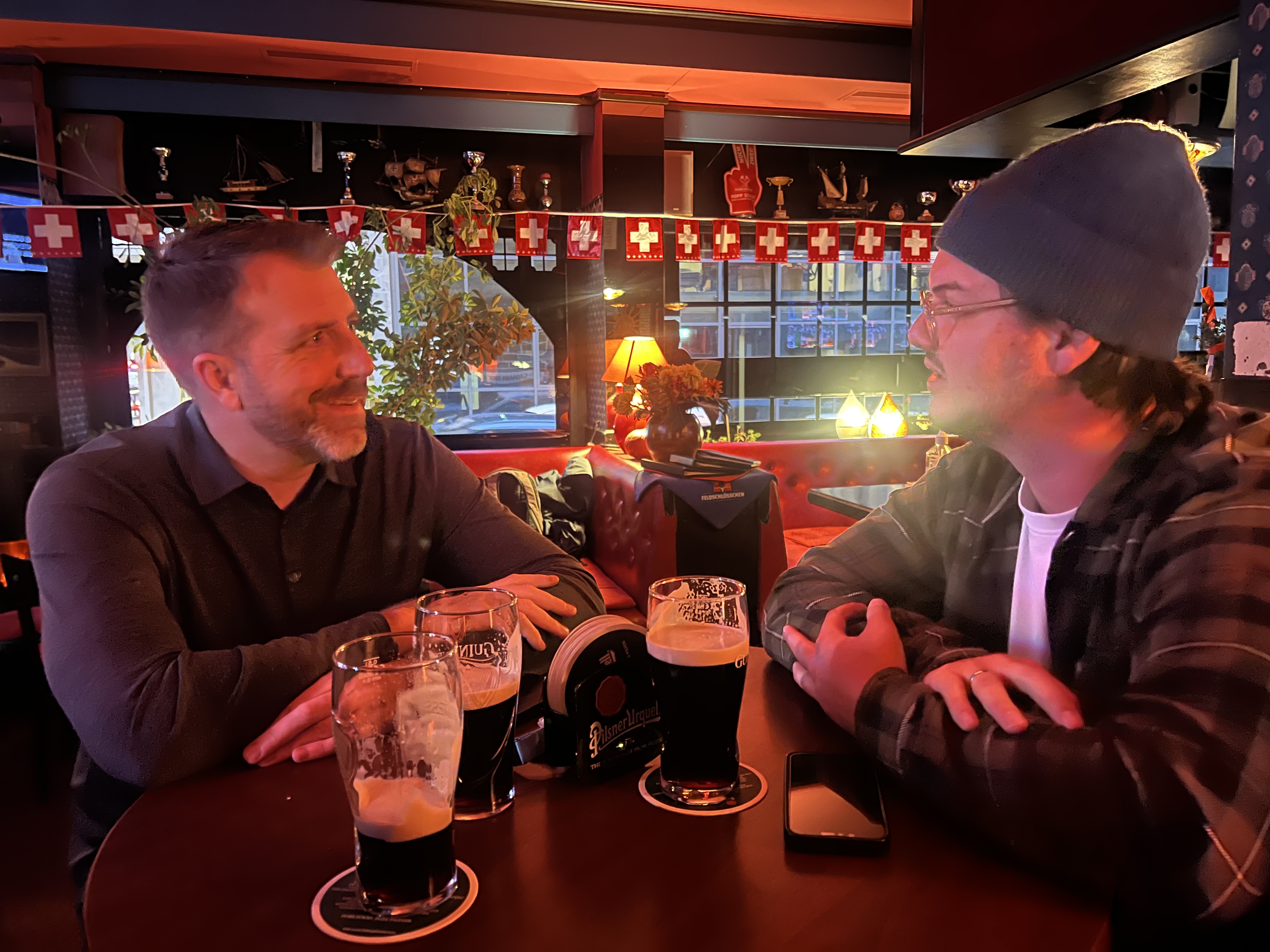 Pub Lord Jim interior in Geneva — two men chatting in a red booth, Swiss flag bunting, multiple Guinness pints on the table