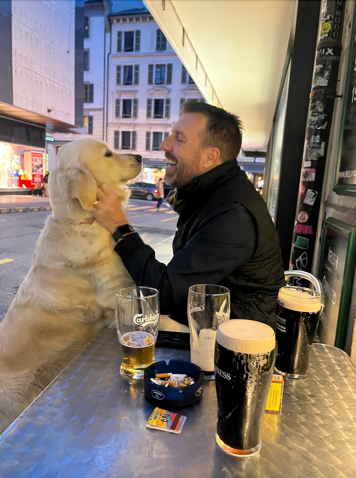 Man laughing with golden retriever outside Mulligans pub at dusk, Geneva