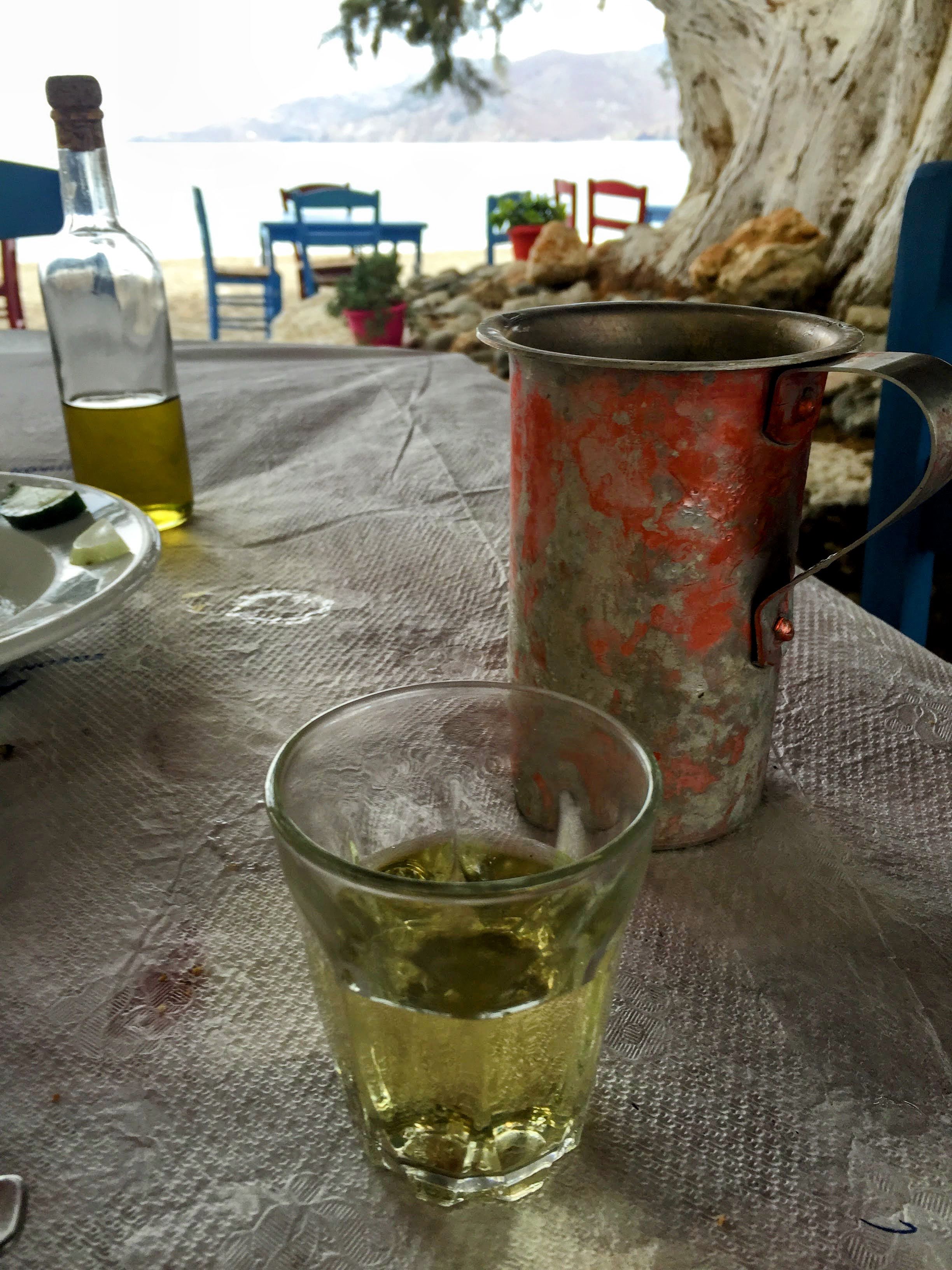 Cold house white wine in a small glass beside a battered metal carafe at a Greek seaside taverna