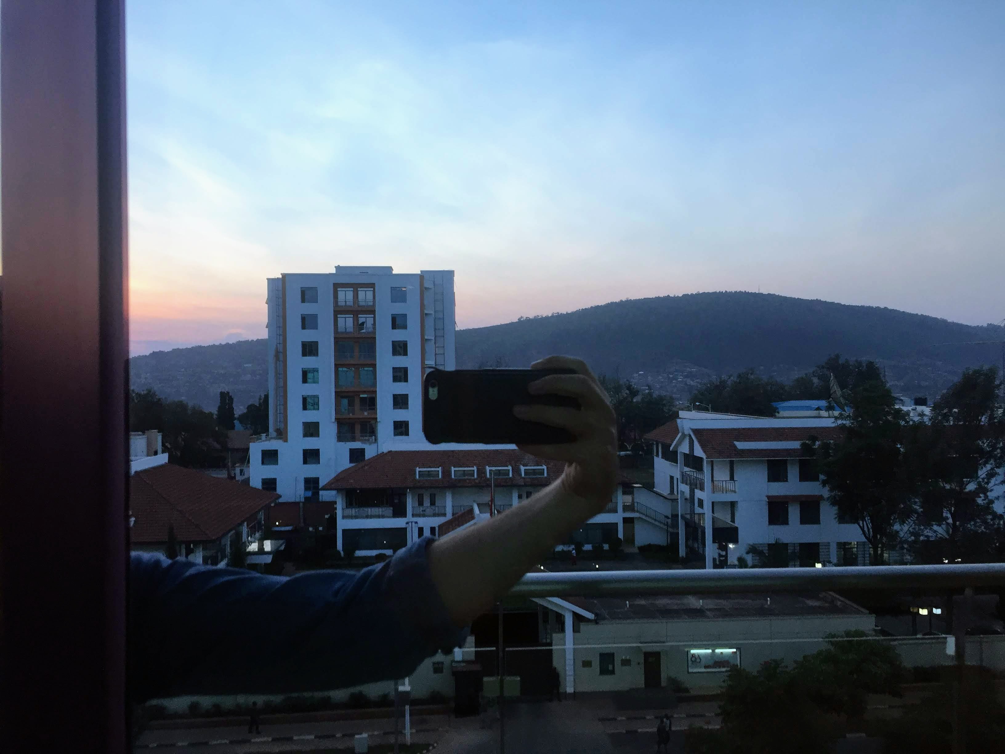 Hand photographing Kigali at dusk from the Marriott window — hills, city lights, sunset