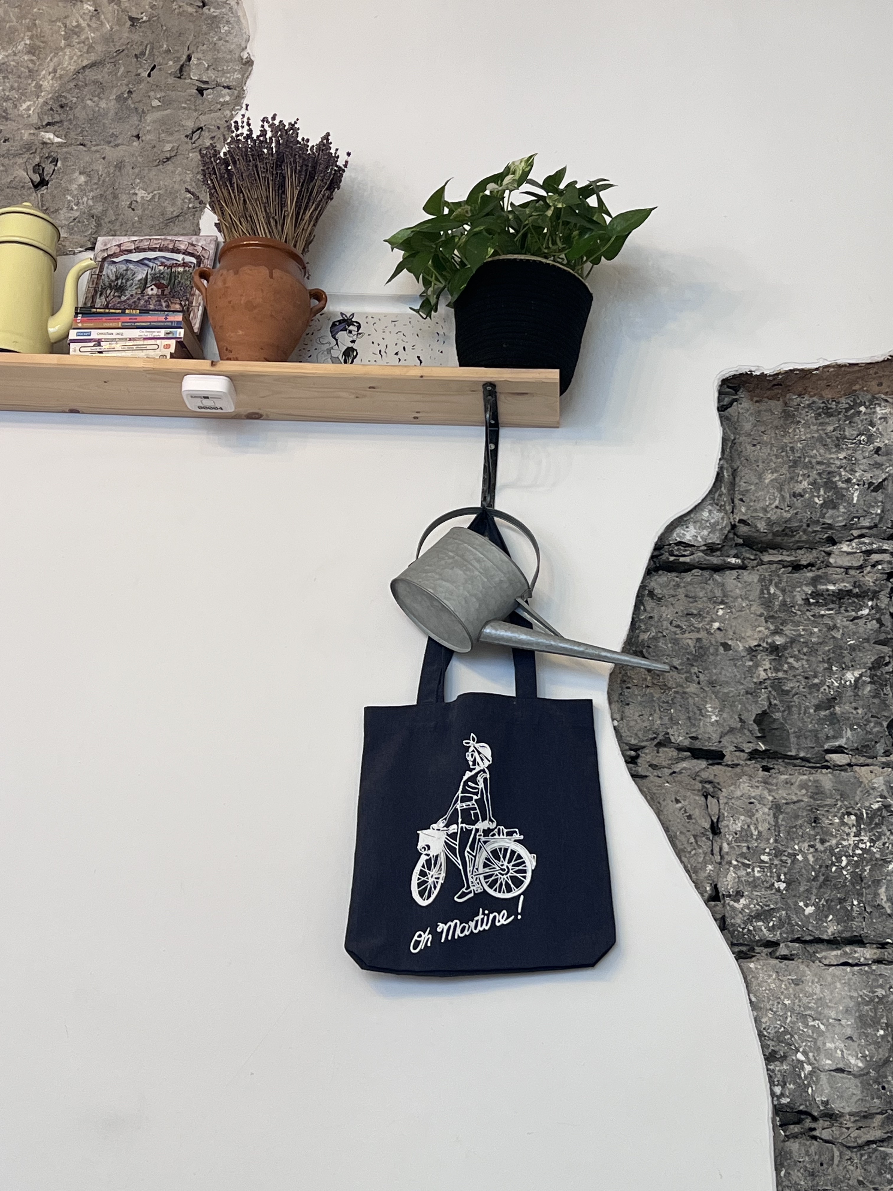 Oh Martine! tote bag hanging on wall — old stone wall, shelf with plants and books