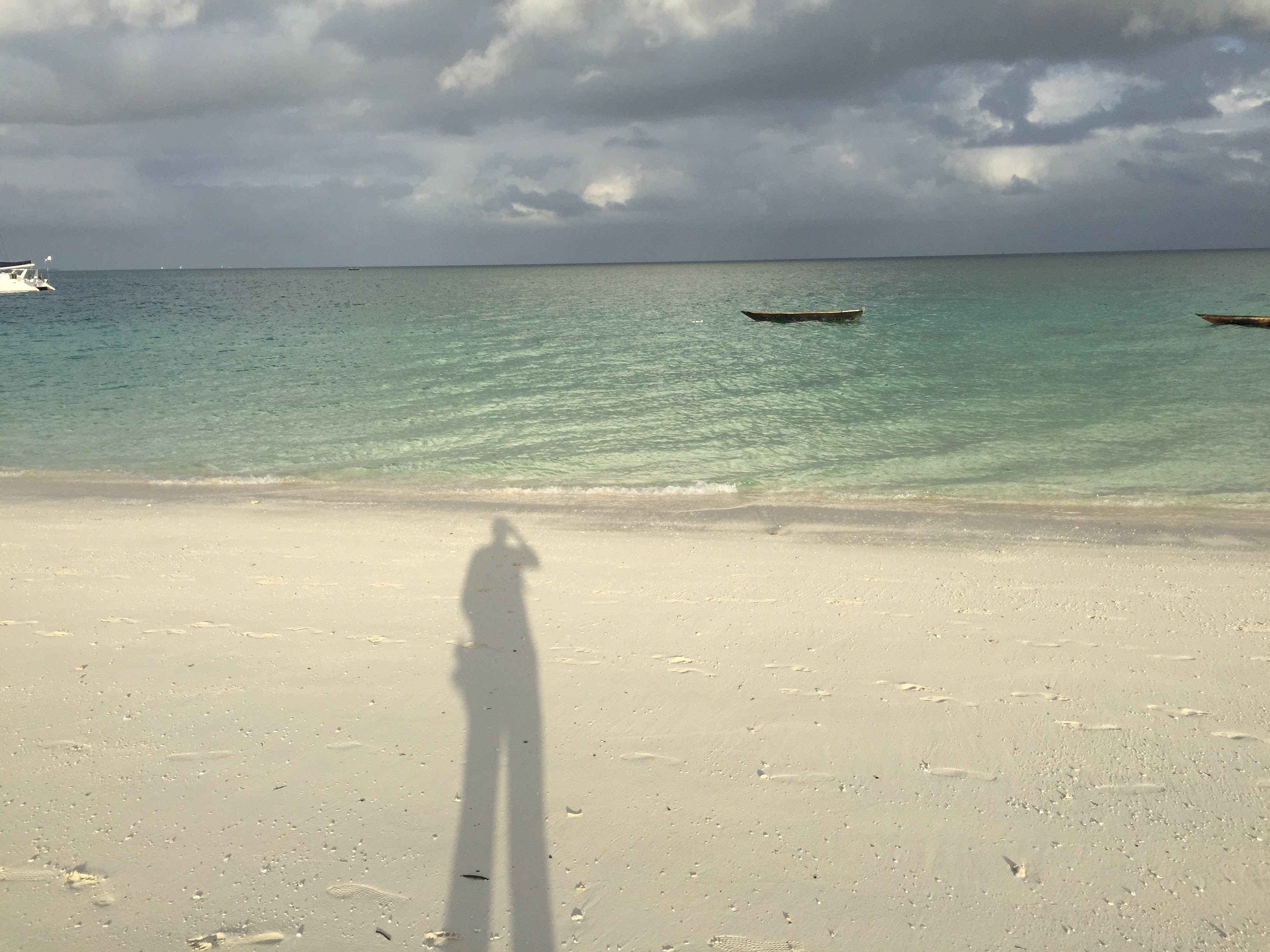 Long shadow of a photographer cast across white sand, dhow boats moored in the turquoise channel