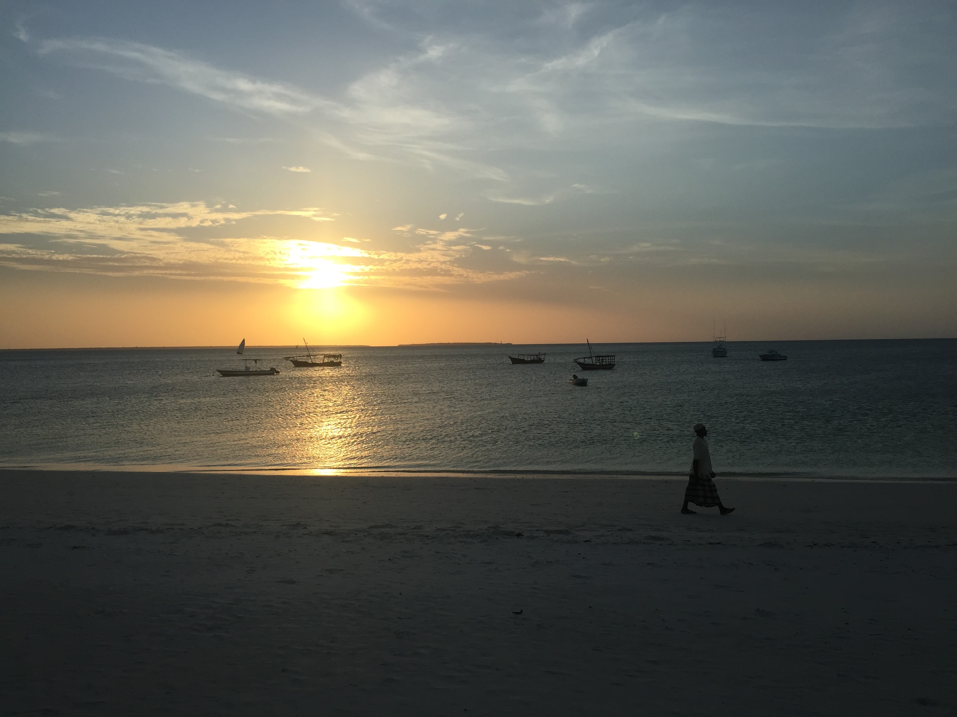 Silhouette of a lone figure walking on the beach at dusk, dhow boats on the horizon, Zanzibar